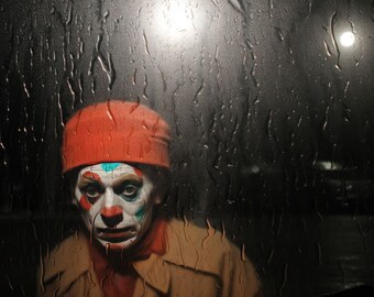 Op de afbeelding: Een clown met een rode hoed en een geschilderd gezicht kijkt uit achter een regenbevuild raam. Het gezicht van de clown is geschilderd met wit, blauw en groen.