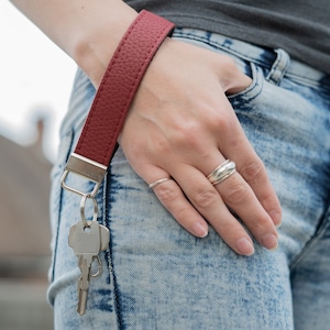 May include: A red leather key chain with a silver key ring and keys attached. The key chain is worn on a person's wrist.