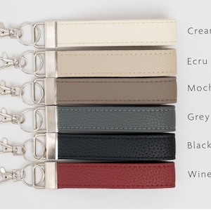 May include: Six leather keychains in different colors: cream, ecru, mocha, grey, black, and wine. Each keychain has a silver key ring.
