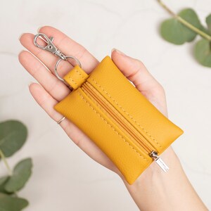 Puede incluir: Un monedero rectangular de color amarillo mostaza con superficie texturizada y cierre de cremallera a juego. El monedero cuenta con un mosquet&oacute;n de metal plateado unido a una anilla, conectada al borde superior por una peque&ntilde;a trabilla.