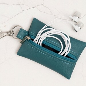 Puede incluir: Un estuche rectangular de color verde azulado con cremallera y superficie texturizada, que cuenta con un mosquet&oacute;n de metal plateado unido a una anilla. El estuche est&aacute; entreabierto, dejando ver un par de auriculares blancos con cable.