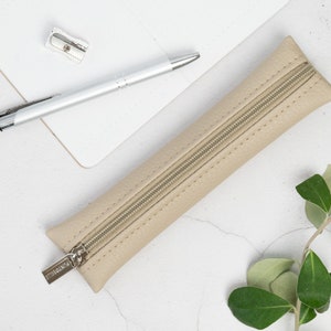 Puede incluir: Un estuche para lápices de cuero beige con cierre de cremallera y un tirador de cremallera plateado. El estuche está sobre una superficie blanca con un bolígrafo y un sacapuntas.