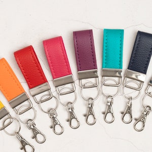 Pode incluir: Uma coleção de oito porta-chaves de couro nas cores amarelo, laranja, vermelho, rosa, roxo, turquesa, azul-marinho e verde-escuro. Cada porta-chaves possui uma tira de couro texturizado, um fecho de metal prateado e uma argola para chaves.