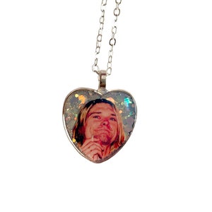 Può includere: Una collana con catena d'argento con un ciondolo a forma di cuore che presenta una foto di un uomo con una camicia rossa e uno sfondo blu chiaro. Il ciondolo a forma di cuore è pieno di glitter iridescente.