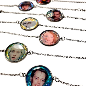 Op de afbeelding: Een collectie zilverkleurige metalen kettingarmbanden, elk voorzien van een ronde hanger met een in hars gegoten portret van een beroemde muzikant of acteur. De portretten zijn omringd door kleurrijke, glinsterende glitterdeeltjes in de hars.