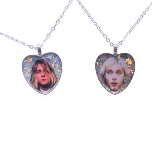 Puede incluir: Dos colgantes en forma de corazón con cadenas plateadas. Cada colgante presenta un retrato de una persona con cabello largo. Los colgantes en forma de corazón están llenos de purpurina y piezas iridiscentes. Las cadenas son delicadas y de color plateado.