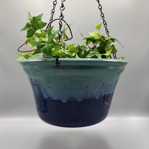 May include: A ceramic hanging planter with a glossy glaze that transitions from a deep navy blue at the base to a lighter teal green at the rim. The planter is suspended by a black metal chain and contains a green ivy plant with pointed leaves.