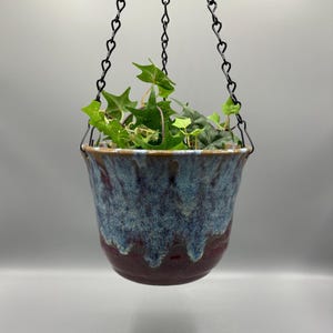 May include: A ceramic hanging planter with a glossy glaze featuring a deep burgundy base that transitions into a textured, drippy blue and white upper section. The pot is suspended by three black metal chains holding a green ivy plant.
