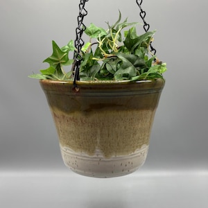 May include: A ceramic hanging planter with a tapered shape, featuring a gradient glaze that transitions from a dark brown rim to a speckled tan middle and a solid off-white base. The planter is suspended by a black metal chain.