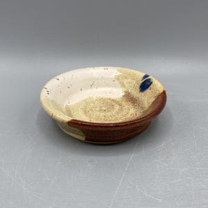 May include: A small, white ceramic bowl with brown and blue glaze. The bowl has a speckled texture and a rounded shape.