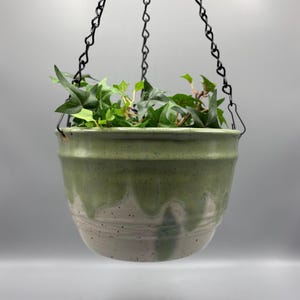 May include: A ceramic hanging planter with a speckled off-white base and a glossy sage green drip glaze finish. The bowl-shaped pot is suspended by three thin black metal chains that meet at a central hook, containing a green ivy plant.