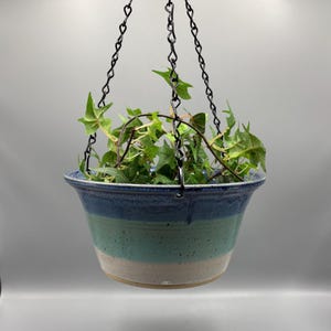 May include: A ceramic hanging planter with a flared rim, featuring a tri-color glaze of dark blue, teal, and off-white with speckled details. The bowl is suspended by three black metal chains attached to the rim, containing a green ivy plant.
