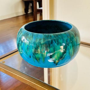 May include: A round, ceramic bowl with a glossy finish. The bowl features a vibrant color palette of turquoise, teal, and green, with a dark blue interior. The bowl is displayed on a glass surface with a gold-colored metal frame.