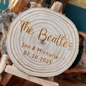 Puede incluir: Una rodaja redonda de madera con el texto "The Beatles" grabado en letras doradas. Debajo del texto se encuentra "Jon & Michelle" y la fecha "07.20.2025" también grabada en oro.