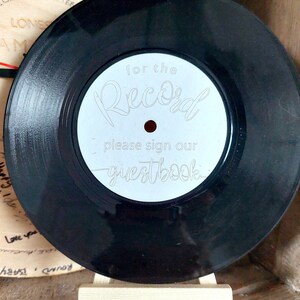Könnte beinhalten: Schwarze Schallplatte mit weißem Label, auf dem "for the Record please sign our guestbook" steht. Die Platte steht auf einer kleinen Holzstaffelei, vermutlich als Gästebuch für eine Veranstaltung.