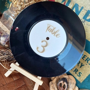 May include: A black vinyl record with a white label that says "Table 3" in gold lettering. The record is on a small wooden easel.