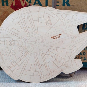 May include: A wooden cutout of the Millennium Falcon spaceship from Star Wars. The cutout is detailed with lines and circles, and has the text "MISHA & IAN 11.05.23" inscribed on it.