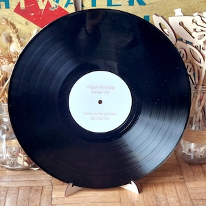 Puede incluir: Disco de vinilo negro con una etiqueta blanca que dice "Happy Birthday Sweet 16!" y "Samantha James 06/06/24". El disco está en un soporte de madera.