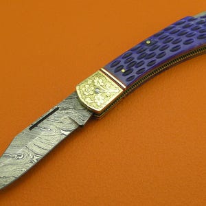 May include: A folding knife with a patterned blade and a purple handle. The handle has a textured design and is accented with gold-colored metal. The blade has a unique pattern. The knife is set against an orange background.