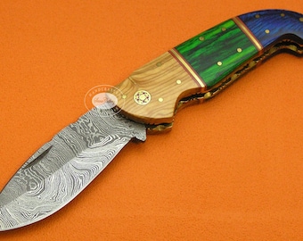 May include: A folding knife with a patterned steel blade and a handle featuring wood, green, and blue accents. The handle is adorned with brass rivets and gold-colored metal accents. The knife is set against an orange background.
