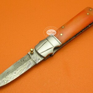 May include: A small, orange-handled folding knife with a Damascus steel blade. The knife has a decorative, silver-colored metal handle with a small, gold-colored button on the side. The knife is handcrafted and has a small, gold-colored loop at the top of the handle.