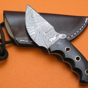 May include: A black handled knife with a Damascus steel blade and a leather sheath. The knife has a lanyard attached to the handle.