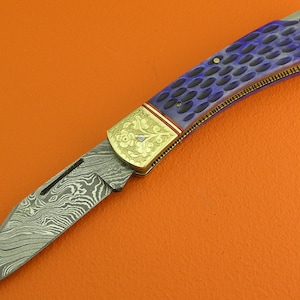 May include: A folding pocket knife with a patterned blade and a blue and black handle. The handle has a decorative gold-colored bolster and end cap. The blade has a unique, swirling pattern. The knife is set against an orange background.