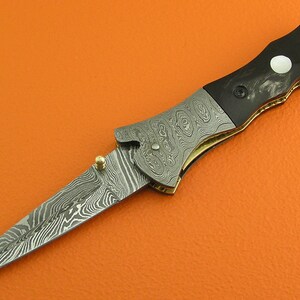 May include: A black and silver folding knife with a patterned blade and a black handle with a white inlay. The knife is open and the blade is extended.