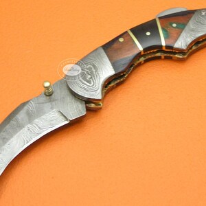 May include: A curved folding knife with a patterned steel blade and a wooden handle. The handle features dark wood, brass accents, and decorative inlays. The knife is open, displaying the blade's sharp edge.