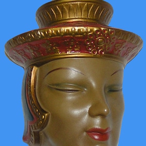 May include: A ceramic wall sculpture of a woman's face with closed eyes. She is wearing a gold and red crown with intricate details and a gold and blue necklace. The sculpture is painted in shades of brown, gold, red, and blue.