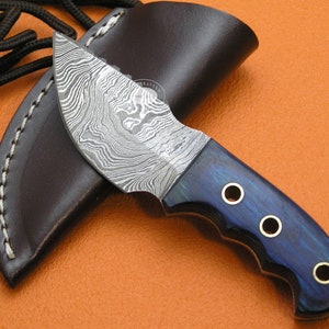 Puede incluir: Un cuchillo pequeño y curvo con una hoja de acero damasco y un mango de madera azul. El mango tiene tres detalles de latón. El cuchillo está en una funda de cuero marrón.