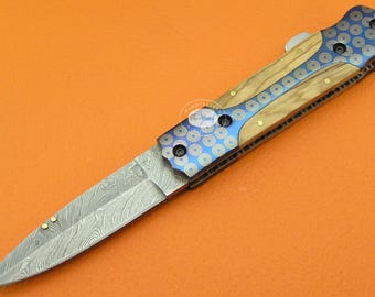 May include: A folding knife with a patterned blade and a handle featuring wood and blue metal accents. The blue metal is decorated with a circular pattern. The knife has a silver blade with a unique design.