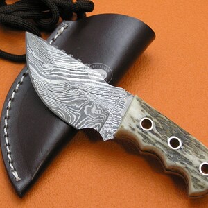 May include: A small, handcrafted knife with a damascus steel blade and a stag horn handle. The knife has a leather sheath with a stitched edge and a hand-stitched loop.