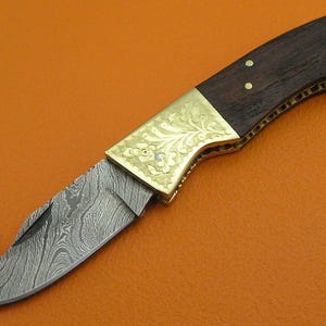 May include: A folding knife with a Damascus steel blade and a dark wood handle. The handle is accented with brass and decorative floral engravings. The blade has a curved, hook-like tip. The knife is set against an orange background.