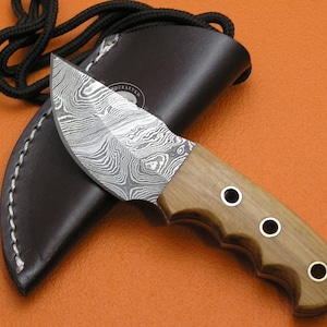 Puede incluir: Un cuchillo pequeño y curvo con una hoja de acero estampada y un mango de madera con detalles de latón. El cuchillo está en una funda de cuero marrón con detalles de costura blanca.