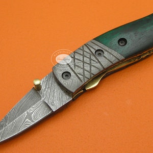 May include: A Damascus steel folding knife with a green wooden handle and brass accents. The blade has a unique pattern, and the handle is secured with black screws. The knife is shown against an orange background.
