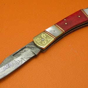 May include: A folding knife with a patterned blade and a red handle. The handle has gold-colored accents and white inlays. The knife is open, displaying the blade's intricate design. The knife is set against an orange background.