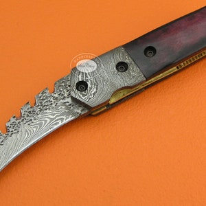 May include: A Damascus steel folding knife with a curved blade and a dark wood handle. The handle has a brass accent and a decorative pattern.