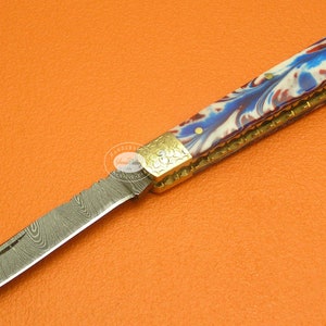 Puede incluir: Cuchillo plegable de acero de Damasco con mango veteado en tonos azules, rojos y blancos. El mango está adornado con metal dorado y remaches de latón. La hoja presenta un patrón distintivo.