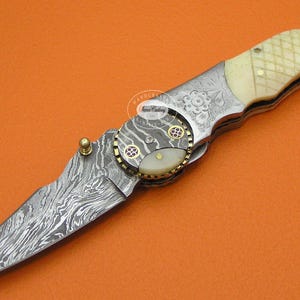 May include: A detailed folding knife with a Damascus steel blade and a cream-colored handle. The handle features a decorative pattern and is accented with brass and a white oval inlay. The knife is set against an orange background.