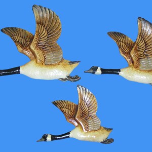 May include: Three ceramic geese in flight, set against a blue background. The geese have detailed brown and tan wings, black necks, and white and tan bodies. The wings are spread, suggesting motion.