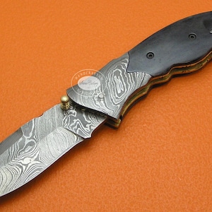 May include: A folding knife with a patterned blade and a dark wooden handle. The blade has a unique swirling pattern, and the handle is secured with brass rivets. The knife is designed for everyday carry and outdoor use.
