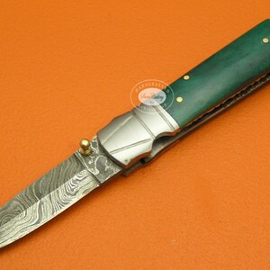 May include: A green and silver folding knife with a patterned blade and a gold accent. The handle is made of green wood with gold accents.