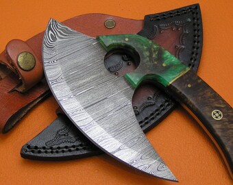 Op de afbeelding: Een Damascus stalen cleaver met een groene en bruine handgreep, rustend op een bruine leren schede. Het mes heeft een unieke gebogen vorm en een kenmerkend patroon. De handgreep is gemaakt van hout en groene hars.