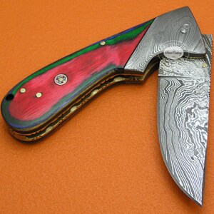 May include: A folding pocket knife with a red, green, and blue wood handle and a Damascus steel blade. The blade is partially open and the handle has a brass inlay. The knife is made by "Sana Cutlery USA".
