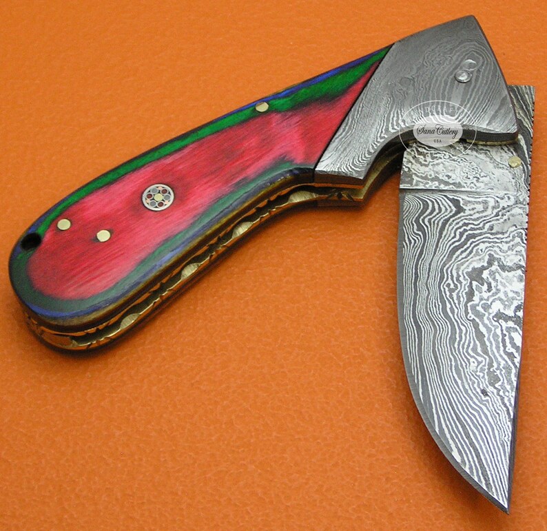 May include: A folding pocket knife with a red, green, and blue wood handle and a Damascus steel blade. The blade is partially open and the handle has a brass inlay. The knife is made by "Sana Cutlery USA".