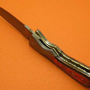 May include: A folding knife with a Damascus steel blade and a wood handle with gold accents.