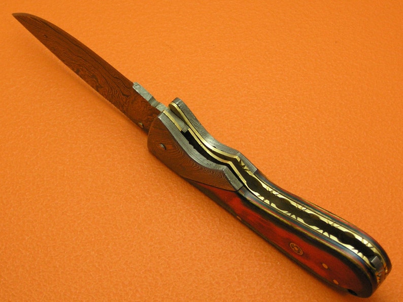May include: A folding knife with a Damascus steel blade and a wood handle with gold accents.