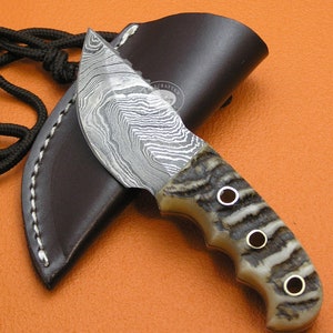 Puede incluir: Un pequeño cuchillo artesanal con una hoja de acero Damasco y una funda de cuero marrón. El mango está hecho de asta de ciervo y tiene un patrón decorativo. El cuchillo mide aproximadamente 18 cm de largo.
