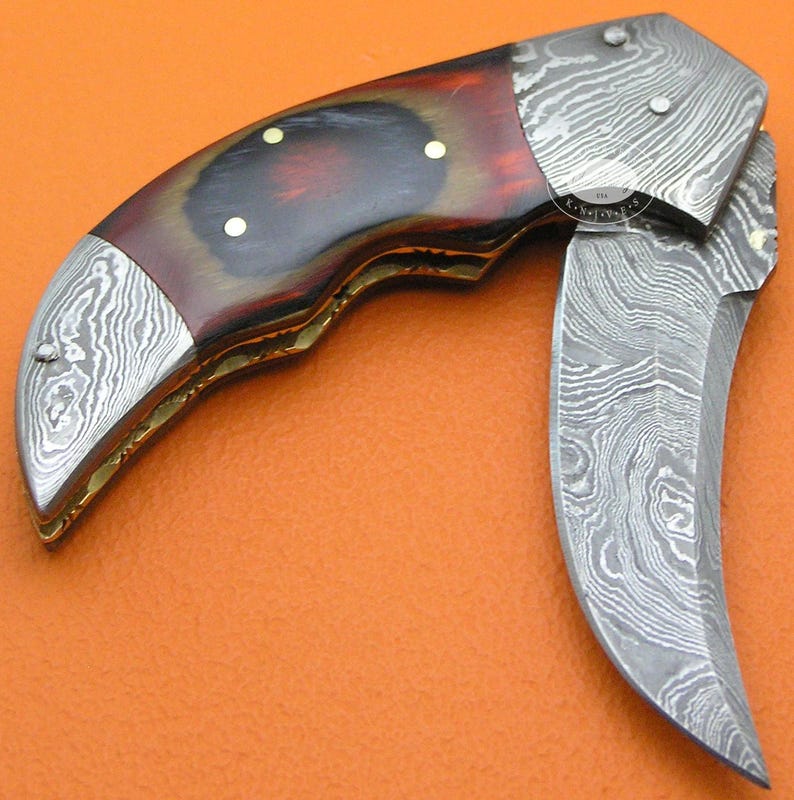 May include: A folding knife with a curved blade and a handle made of wood and metal. The handle has a dark red and brown color scheme with brass rivets. The blade and metal accents feature a patterned design.